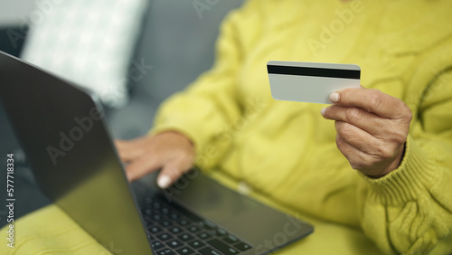 Middle age hispanic woman using laptop and credit card sitting on sofa at home