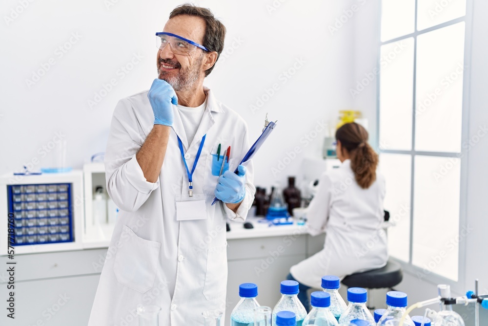 Middle age man working at scientist laboratory serious face thinking about question with hand on chin, thoughtful about confusing idea