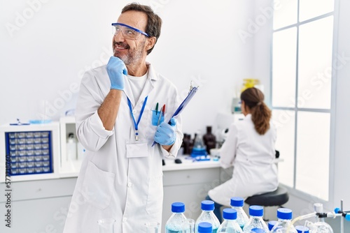 Middle age man working at scientist laboratory serious face thinking about question with hand on chin  thoughtful about confusing idea