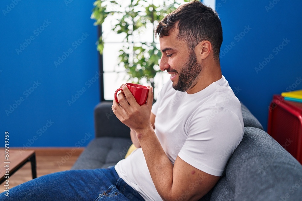 Young hispanic man drinking coffee sitting on sofa at home