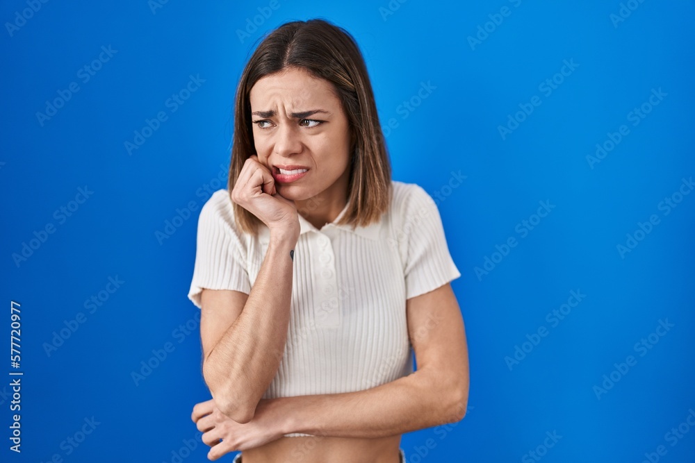 Hispanic woman standing over blue background looking stressed and nervous with hands on mouth biting nails. anxiety problem.