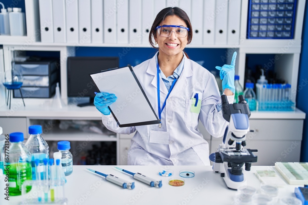 Hispanic young woman working at scientist laboratory pointing finger up with successful idea. exited and happy. number one. Hispanic young woman working at scientist laboratory pointing finger up with successful idea. exited and happy. number one.