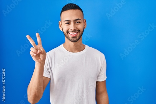 Young hispanic man standing over blue background smiling with happy face winking at the camera doing victory sign with fingers. number two.