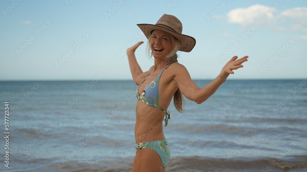 Portrait of smiling happy woman with arms outstretched arms wearing bikini and hat with the blue ocean behind her.