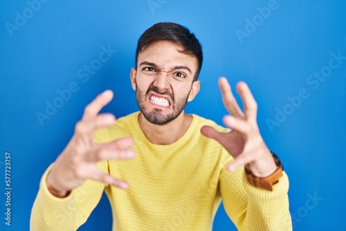 Hispanic man standing over blue background shouting frustrated with rage, hands trying to strangle, yelling mad
