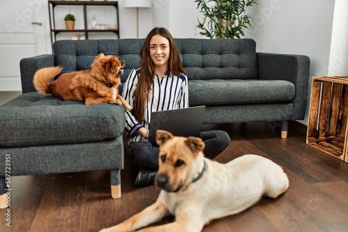 Young hispanic woman using laptop sitting on floor with dogs at home