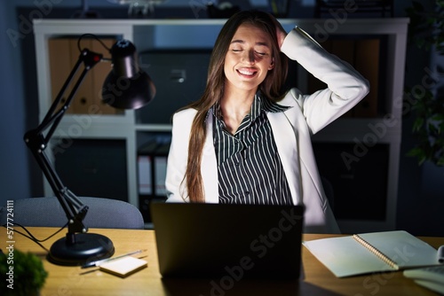Young brunette woman working at the office at night with laptop smiling confident touching hair with hand up gesture, posing attractive and fashionable
