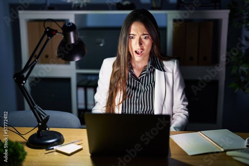 Young brunette woman working at the office at night with laptop in shock face, looking skeptical and sarcastic, surprised with open mouth