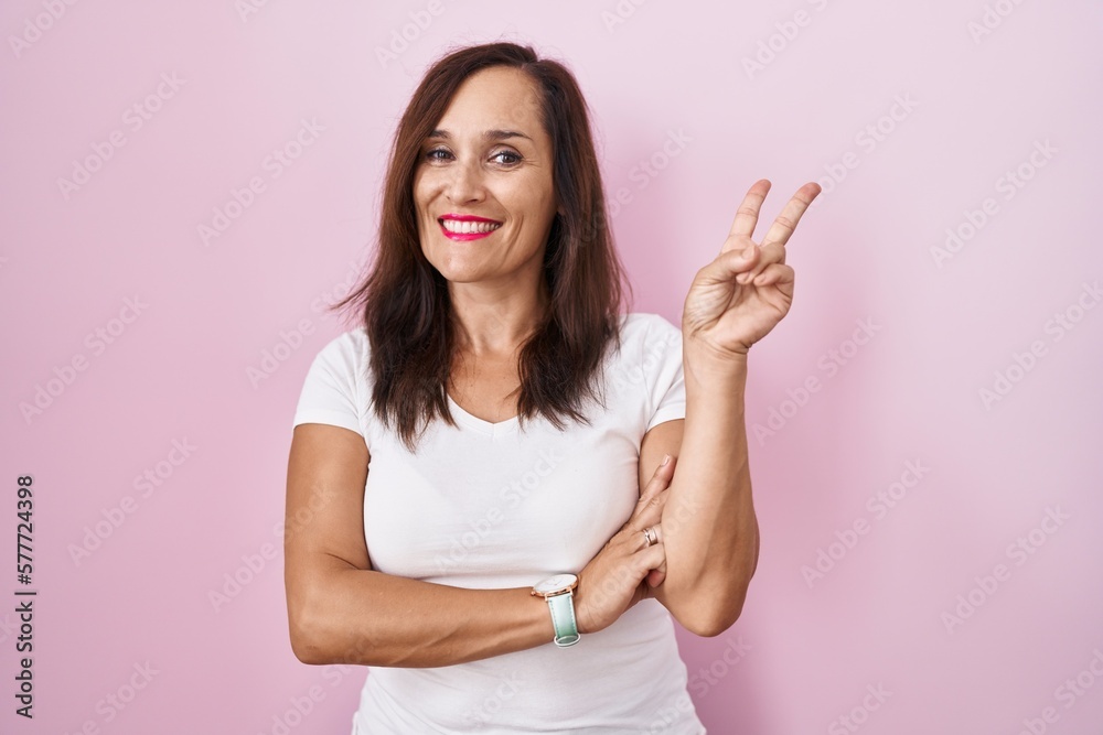 Middle age brunette woman standing over pink background smiling with happy face winking at the camera doing victory sign with fingers. number two. Middle age brunette woman standing over pink background smiling with happy face winking at the camera doing victory sign with fingers. number two.