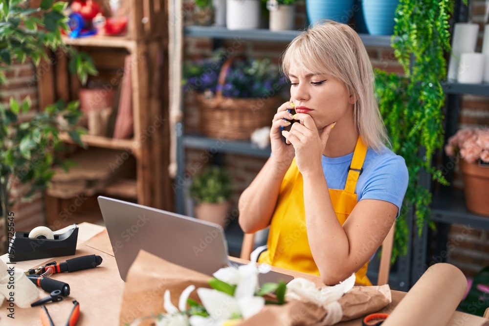 Young blonde woman florist talking on smartphone using laptop at florist