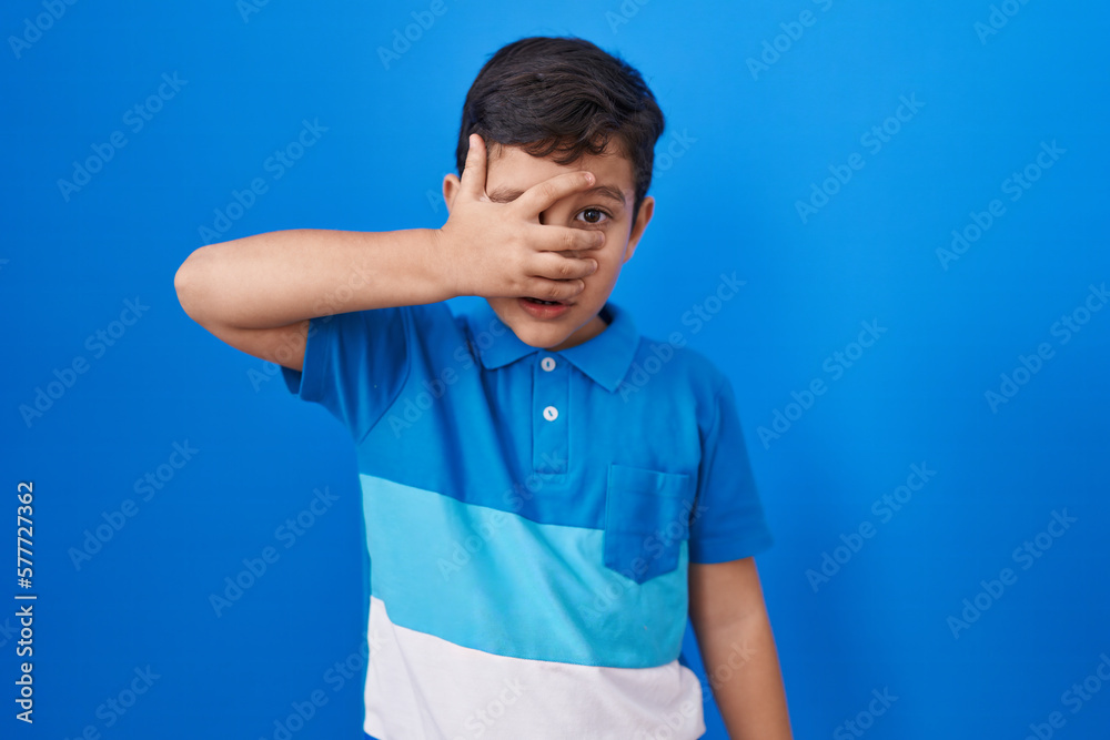 Little hispanic boy standing over blue background peeking in shock covering face and eyes with hand, looking through fingers afraid Little hispanic boy standing over blue background peeking in shock covering face and eyes with hand, looking through fingers afraid
