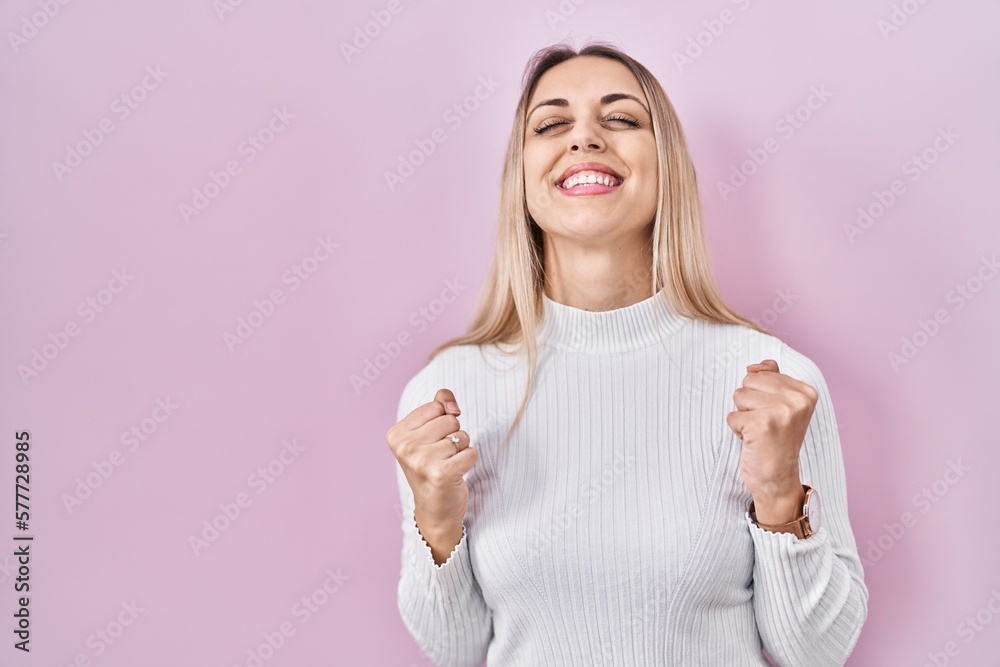 Young blonde woman wearing white sweater over pink background very happy and excited doing winner gesture with arms raised, smiling and screaming for success. celebration concept. Young blonde woman wearing white sweater over pink background very happy and excited doing winner gesture with arms raised, smiling and screaming for success. celebration concept.