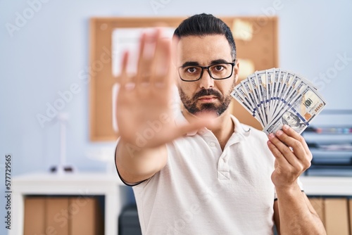 Young hispanic man with beard and glasses holding dollar banknotes with open hand doing stop sign with serious and confident expression, defense gesture