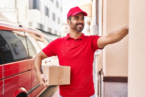 Young hispanic man courier holding package press doorbell at street photo