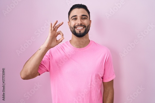 Hispanic young man standing over pink background smiling positive doing ok sign with hand and fingers. successful expression.