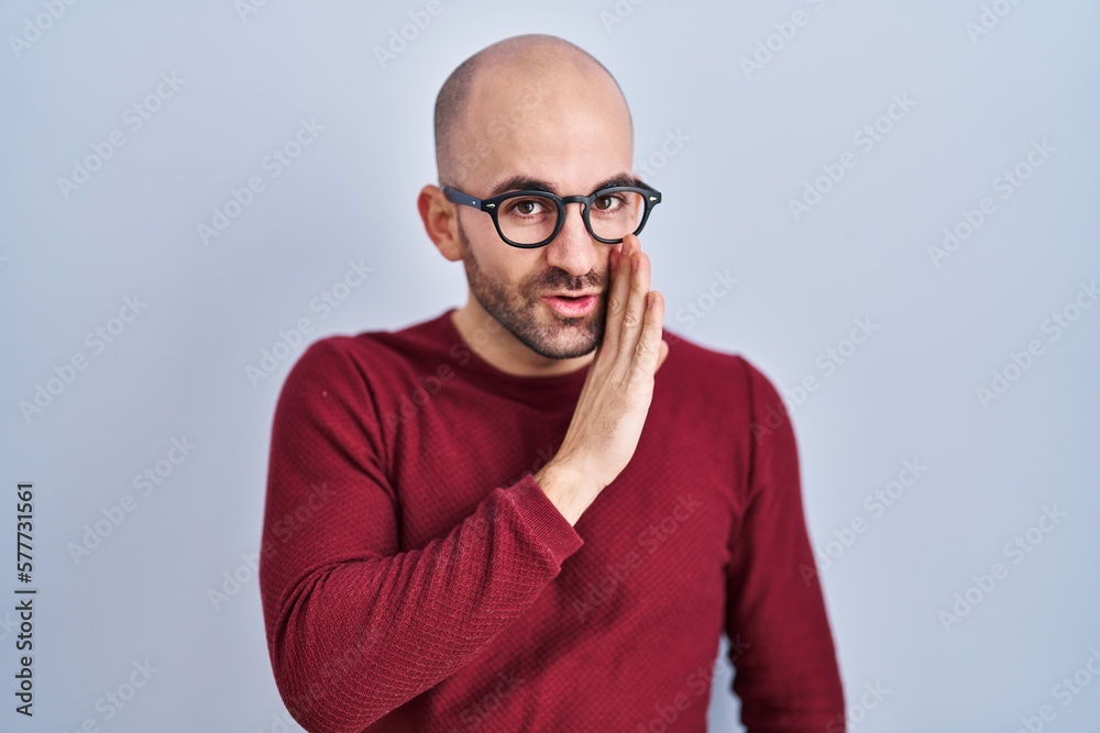Young bald man with beard standing over white background wearing glasses hand on mouth telling secret rumor, whispering malicious talk conversation Young bald man with beard standing over white background wearing glasses hand on mouth telling secret rumor, whispering malicious talk conversation