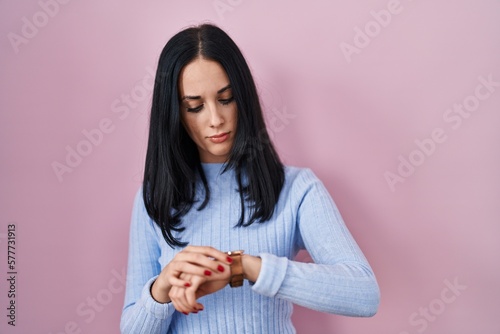 Hispanic woman standing over pink background checking the time on wrist watch, relaxed and confident