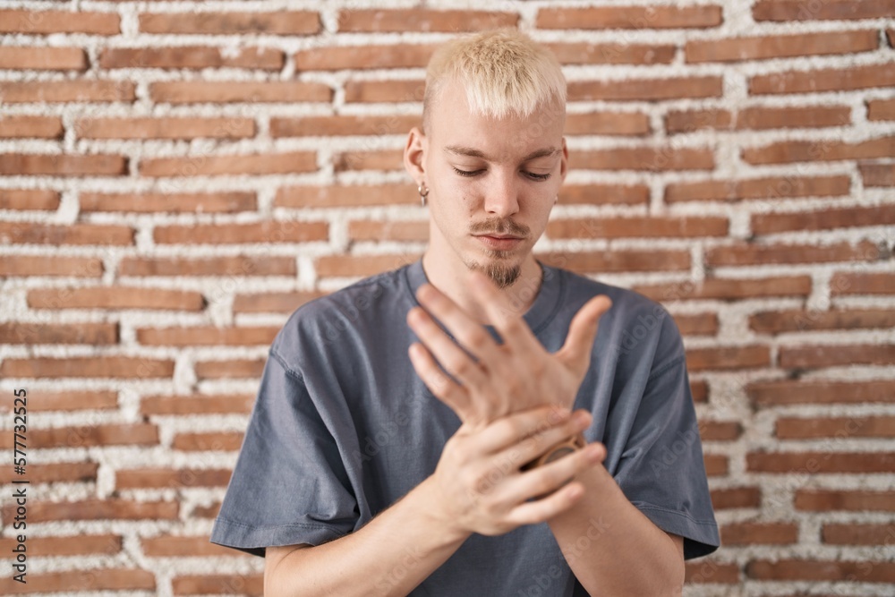 Young caucasian man standing over bricks wall suffering pain on hands and fingers, arthritis inflammation Young caucasian man standing over bricks wall suffering pain on hands and fingers, arthritis inflammation
