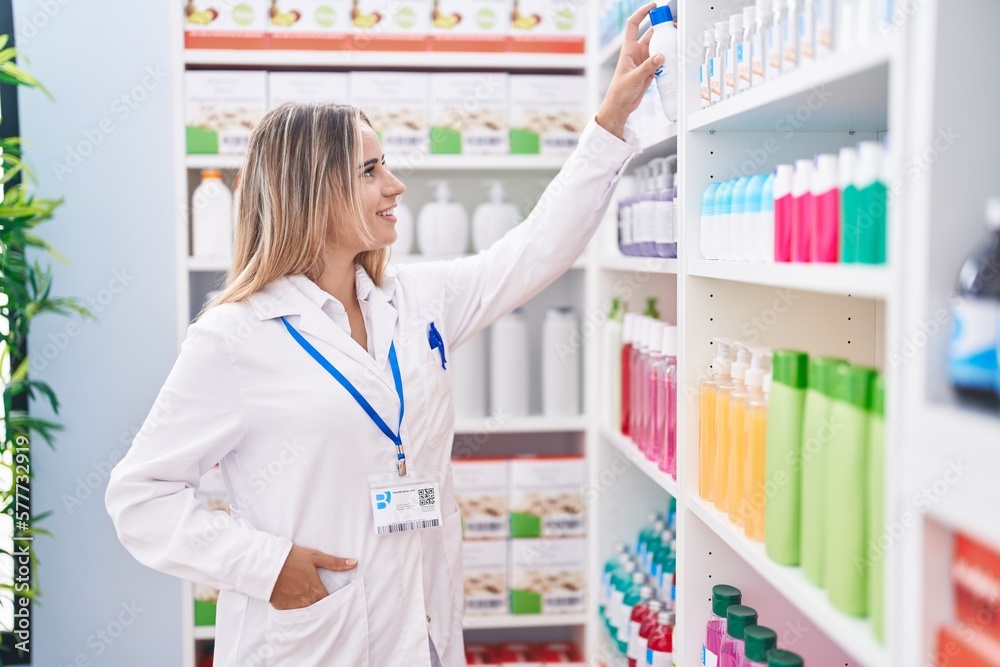Young blonde woman pharmacist smiling confident holding product on shelving at pharmacy