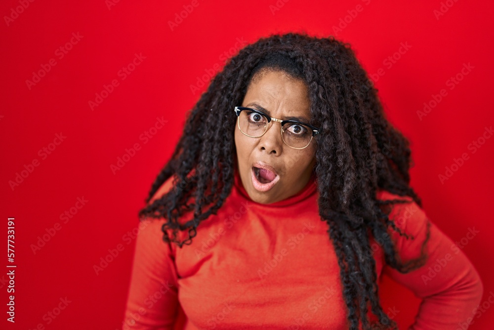 Plus size hispanic woman standing over red background in shock face, looking skeptical and sarcastic, surprised with open mouth