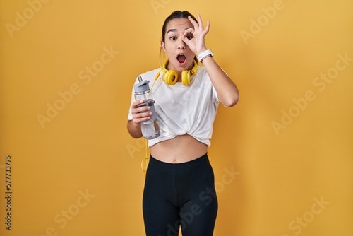 Young south asian woman wearing sportswear drinking water doing ok gesture shocked with surprised face, eye looking through fingers. unbelieving expression.