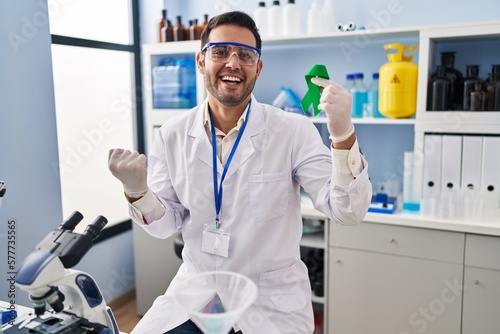 Young hispanic man with beard working at scientist laboratory holding green ribbon screaming proud  celebrating victory and success very excited with raised arms