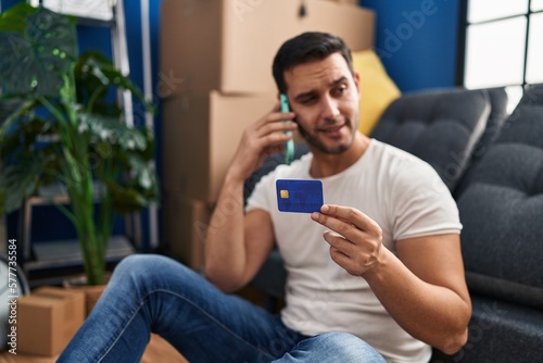 Young hispanic man talking on smartphone using credit card at new home