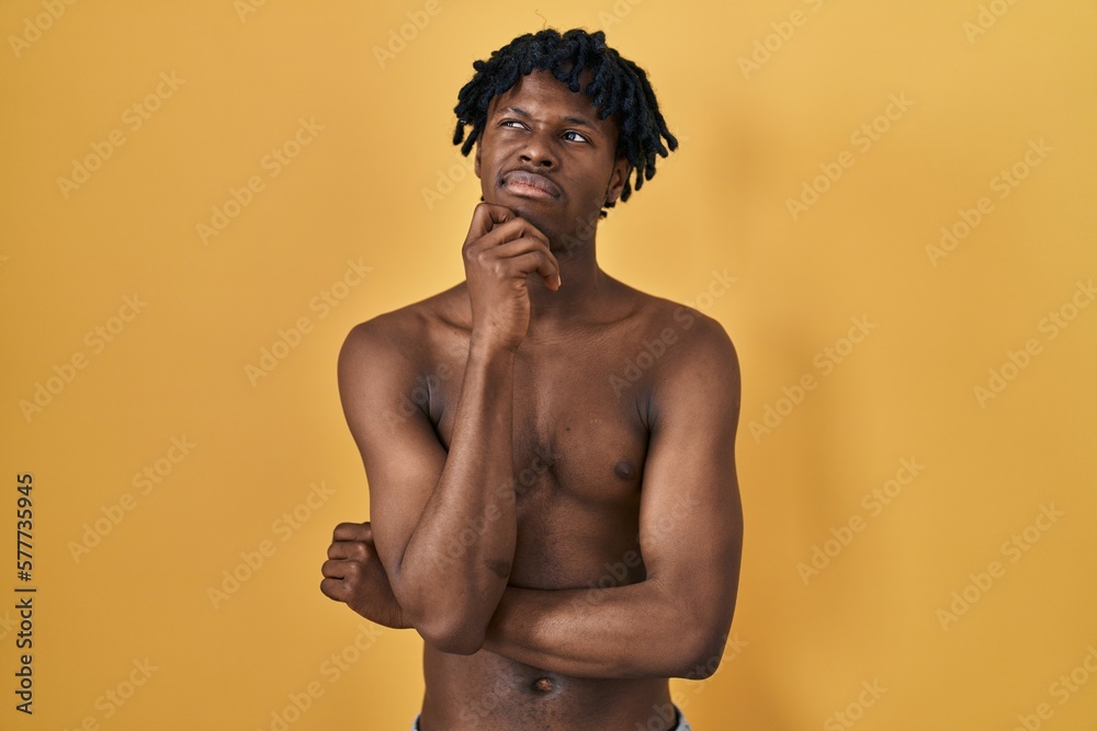 Fototapeta premium Young african man with dreadlocks standing shirtless with hand on chin thinking about question, pensive expression. smiling with thoughtful face. doubt concept.