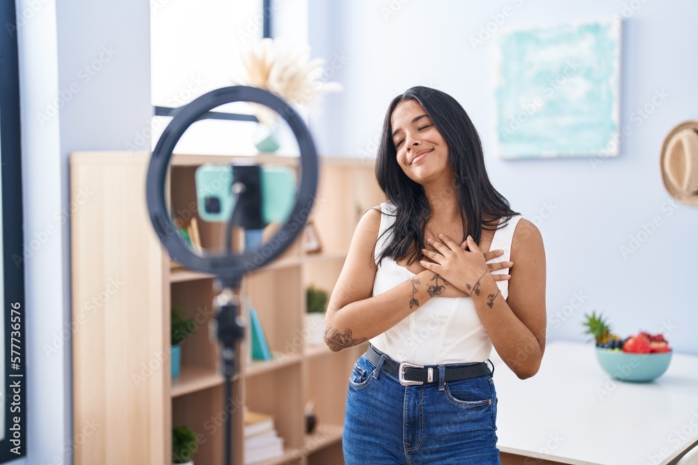 Brunette woman recording vlog tutorial with smartphone at home smiling with hands on chest, eyes closed with grateful gesture on face. health concept.