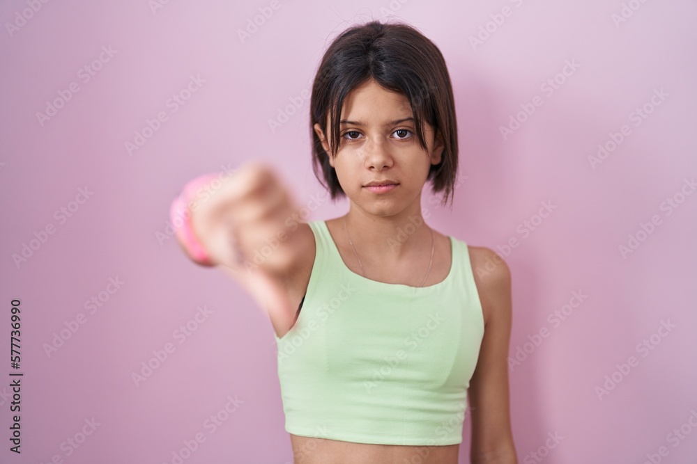 Young girl standing over pink background looking unhappy and angry showing rejection and negative with thumbs down gesture. bad expression. Young girl standing over pink background looking unhappy and angry showing rejection and negative with thumbs down gesture. bad expression.