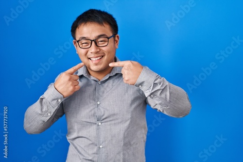 Young chinese man standing over blue background smiling cheerful showing and pointing with fingers teeth and mouth. dental health concept.