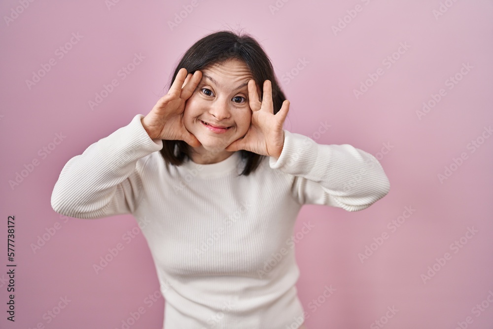 Woman with down syndrome standing over pink background smiling cheerful playing peek a boo with hands showing face. surprised and exited