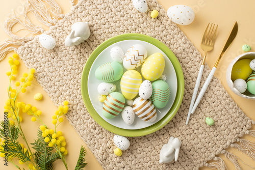 Easter concept. Top view photo of dish with yellow green white easter eggs knife fork ceramic bunnies cloth napkin and mimosa flowers on isolated beige background