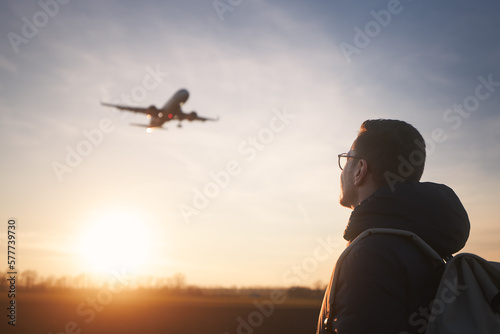 Fotografi Man with backpack looking up to airplane landing at airport during beautiful sunset