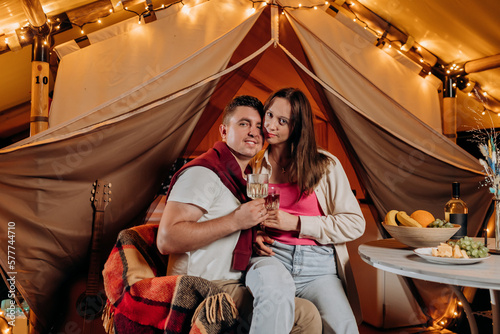 Happy lovely couple relaxing in glamping on summer evening and drinking wine near cozy bonfire. Luxury camping tent for outdoor recreation and recreation. Lifestyle concept © bondvit