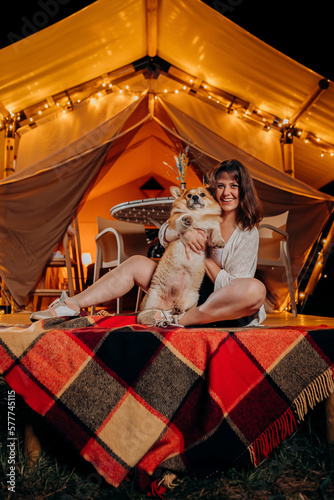 Happy young woman with her Welsh Corgi Pembroke dog relaxing in glamping on summer evening near cozy bonfire. Luxury camping tent for outdoor recreation and recreation. Lifestyle concept