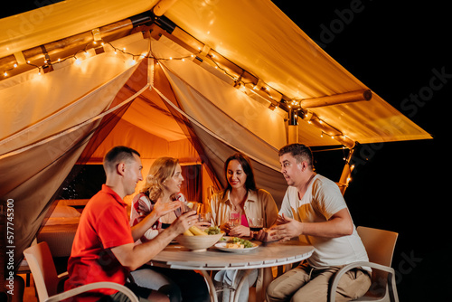 Happy group of friends relaxing in glamping and drinking wine on summer evening near cozy bonfire. Luxury camping tent for outdoor recreation and recreation. Lifestyle concept © bondvit