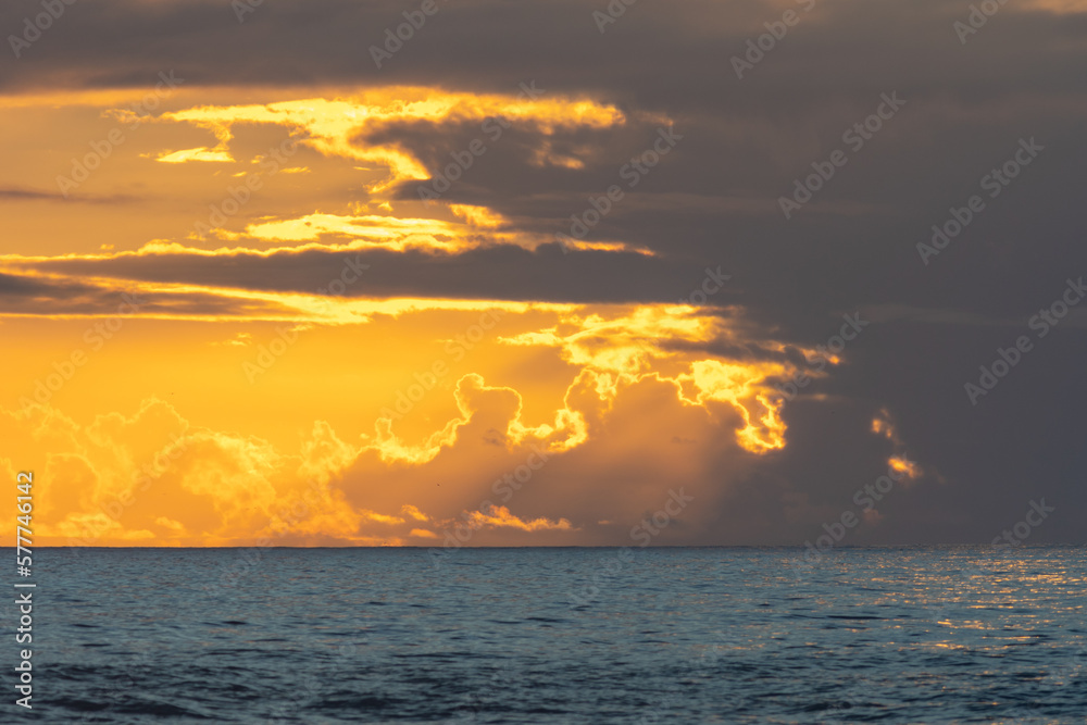 Dark blue sea at yellow sunset and sun breaking through dark gray clouds