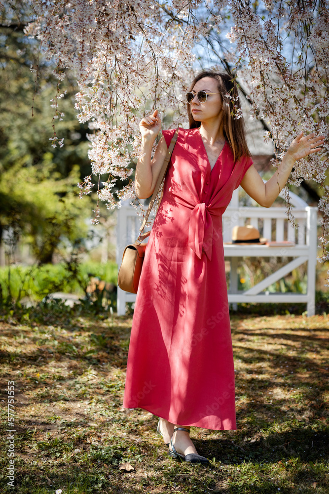 A beautiful young woman walk through a meadow in a blooming spring garden against the backdrop of a white elegant blossom sakura, an exquisite bench A beautiful young woman walk through a meadow in a blooming spring garden against the backdrop of a white elegant blossom sakura, an exquisite bench