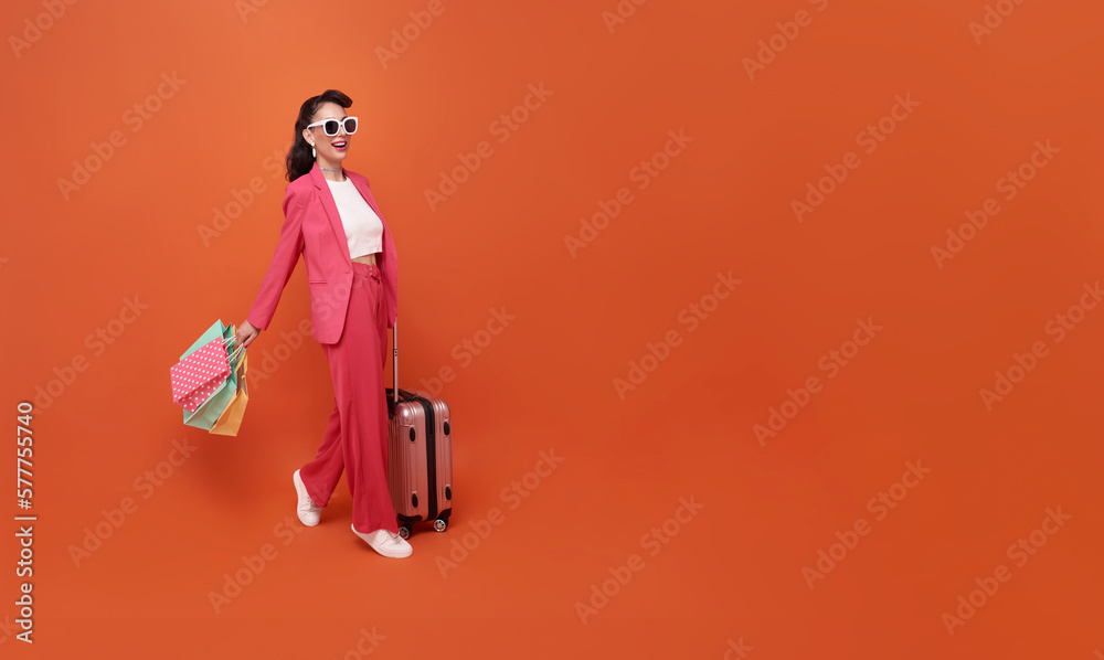 Happy young woman traveler drag luggage and shopping bag isolated on orange copy space ackground, Tourist girl having cheerful holiday trip concept, Full body composition