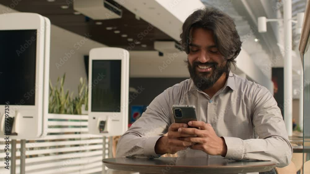 Happy young indian businessman holding smartphone sending message sitting indoors smiling arabian man play mobile game using application on phone reading good news on internet chatting in social media