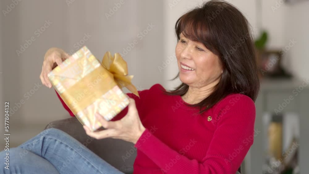 Attractive stylish woman wearing red top smiling, happy holding colorful wrapped birthday present, enjoying turning looking at gift sitting on living room chair. Mature asian female surprised. Attractive stylish woman wearing red top smiling, happy holding colorful wrapped birthday present, enjoying turning looking at gift sitting on living room chair. Mature asian female surprised.