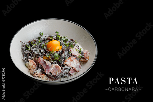 Italian pasta Carbonara with black dough spaghetti, bacon Pancetta, creamy sauce and egg yolk on top. Dish isolated on black background. Ready advertising banner with text space and copy space photo