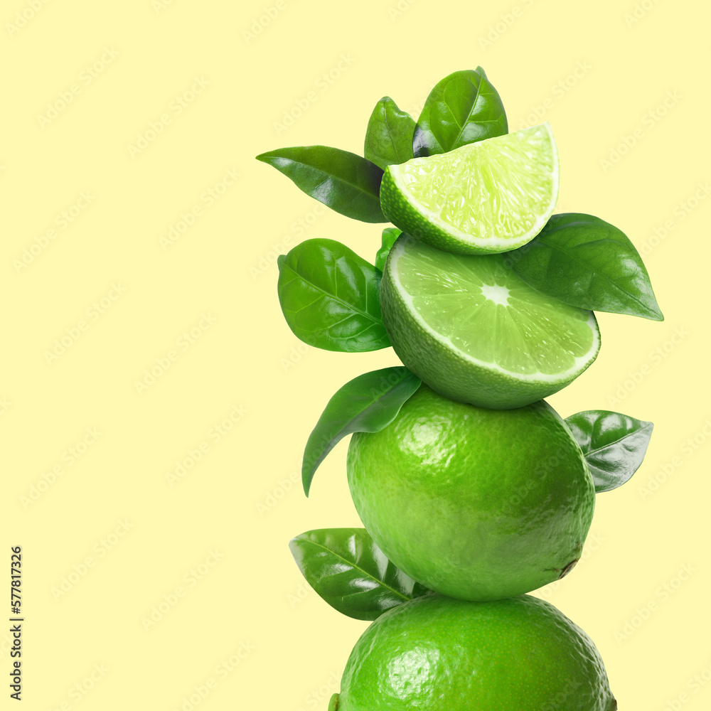 Stacked cut and whole limes with green leaves on pale light yellow background, space for text