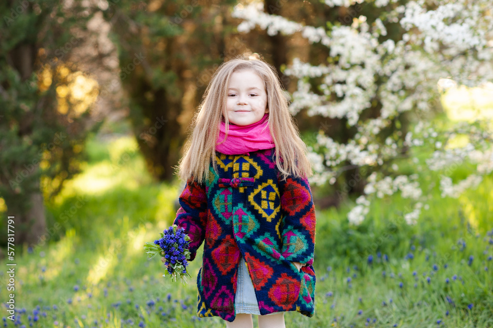 Smiling stylish kid girl 5-6 year old wearing trendy coat holding flowers over nature background outdoor. Looking at camera. Childhood. Spring season. Smiling stylish kid girl 5-6 year old wearing trendy coat holding flowers over nature background outdoor. Looking at camera. Childhood. Spring season.