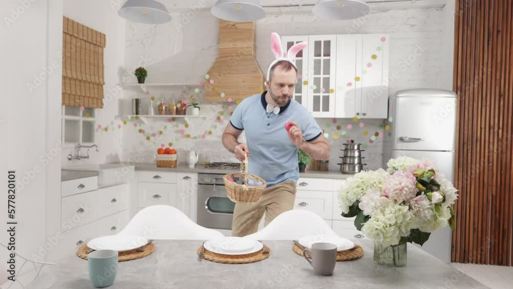 Portrait of a man in fluffy bunny ears hiding colorful eggs in his stylish home. Adult, caucasian parent preparing Easter entertainment for his children. High quality 4k footage Portrait of a man in fluffy bunny ears hiding colorful eggs in his stylish home. Adult, caucasian parent preparing Easter entertainment for his children. High quality 4k footage