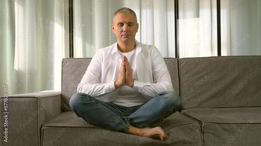 A man sits in a lotus position in a room on a meditation sofa, closes his eyes. The man is doing yoga. A man sits in a lotus position in a room on a meditation sofa, closes his eyes. The man is doing yoga.
