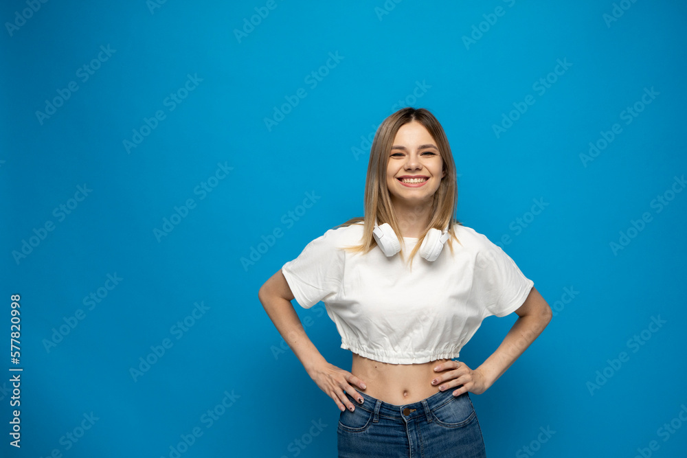 Beautiful young blonde woman with headphones and mobile device listening to music and smiling and dancing, isolated on blue background.