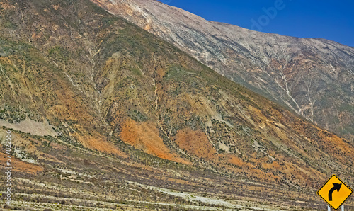 Fototapeta Naklejka Na Ścianę i Meble -  Imposing steep bare colorful mountain wall in desert landscape, blue sky, lonely yellow lost curve right direction sign