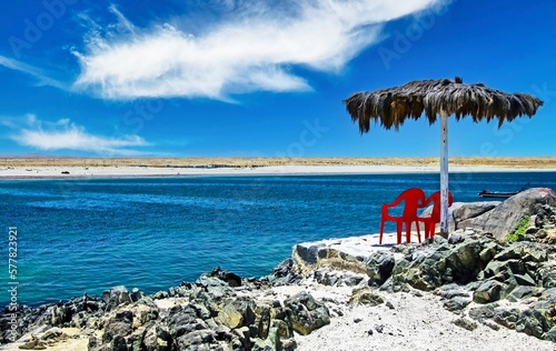 2 lonely empty red chairs under a palm leaf parasol on a stone terrace on a rocky beach on a beautiful sea lagoon with a white sand - Bahia Inglesa, Chile